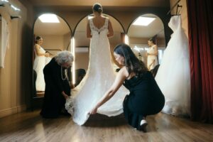 A bride standing in a fitting room while a seamstress adjusts the hem of her wedding gown